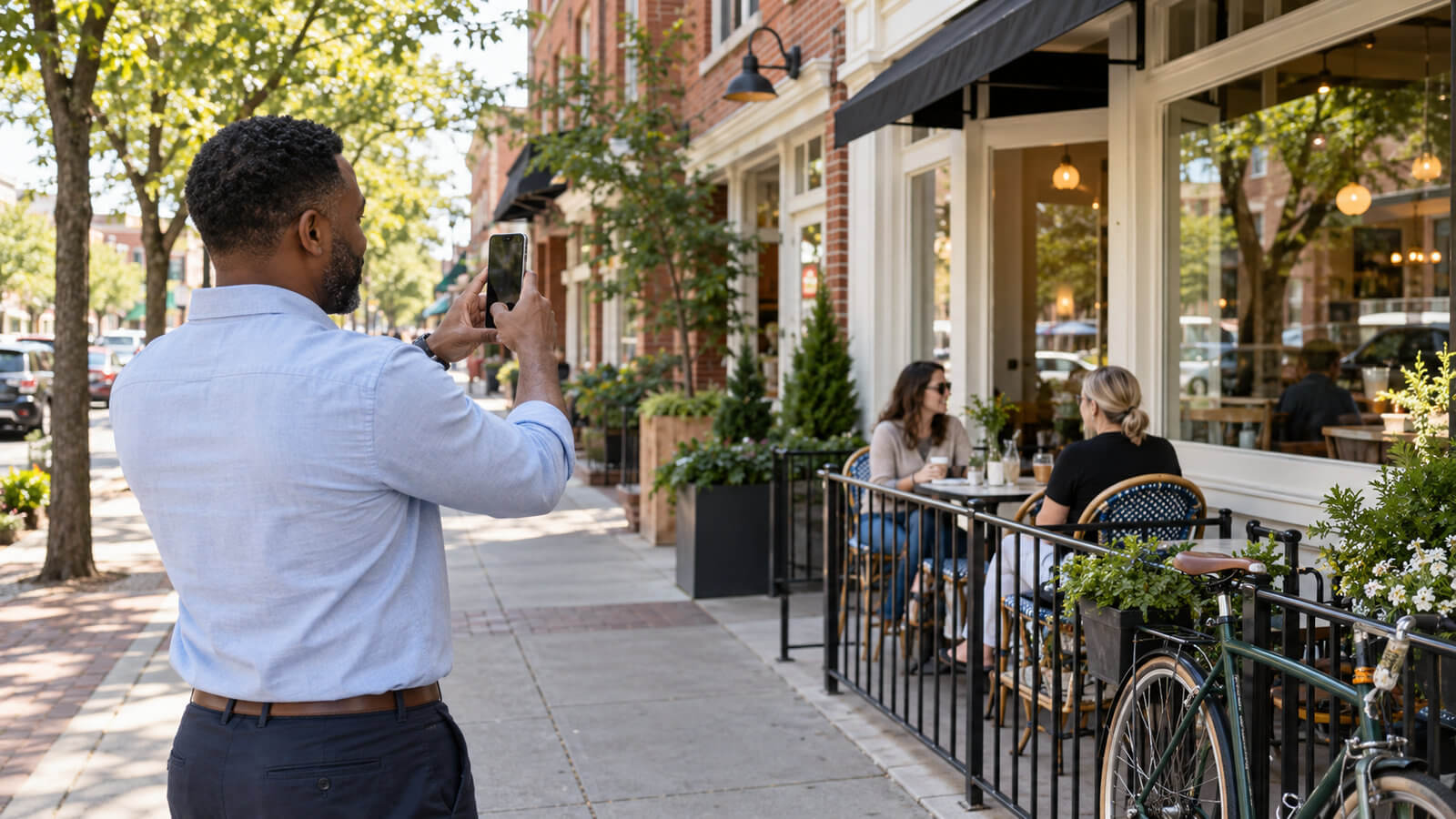 A man photographs a neighborhood café with his smartphone while customers sit on its sidewalk patio.